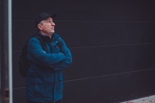 Thoughtful senior man aged 50s 60s in blue jacket and cap standing alone outdoors looking into distance, psychological mental health concept of loneliness and reflection