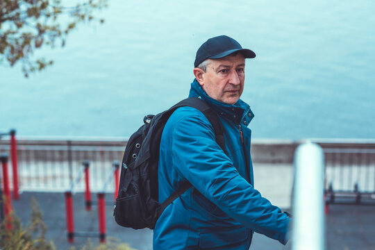 Thoughtful senior man aged 50s 60s in blue jacket and cap standing alone outdoors looking into distance, psychological mental health concept of loneliness and reflection