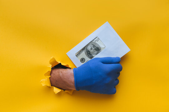 The right male hand in a blue fabric work glove holds an envelope with dollar bills (money). Torn hole in yellow paper. Concept of guest worker, handyman, gray illegal salary for work. Copy space.