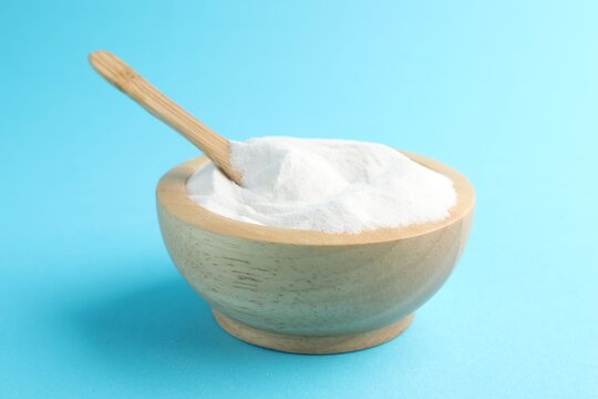 Baking soda and spoon in wooden bowl on light blue background, closeup