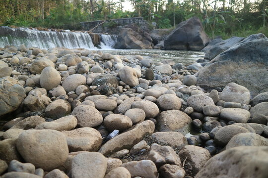 Scattered smooth river stones and rocks on dry riverbed with small waterfall flowing gently in the background surrounded by lush tropical green trees