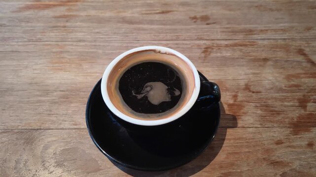 Top view of black coffee in a ceramic cup as crema slowly swirls across the surface on a rustic wooden table.