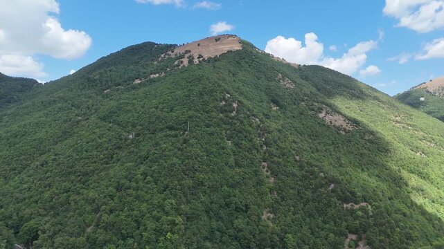 Montagna ricoperta da foresta verde con pendii ripidi, zona rocciosa vicino alla cima e cielo azzurro con nuvole bianche. Paesaggio naturale ampio, luminoso e incontaminato