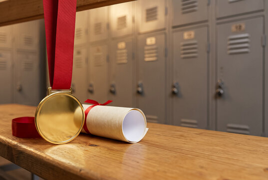 A gold medal on a red ribbon and a rolled-up diploma on a wooden bench in a school locker room with metal lockers.
