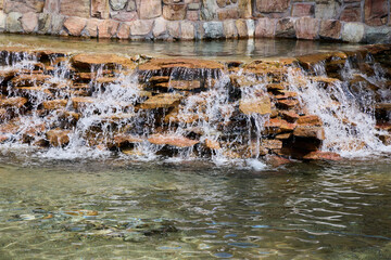 A beautiful artificial waterfall cascading over multi-tiered brown stones into a clear water pond or swimming pool. © beautiful-photo.de