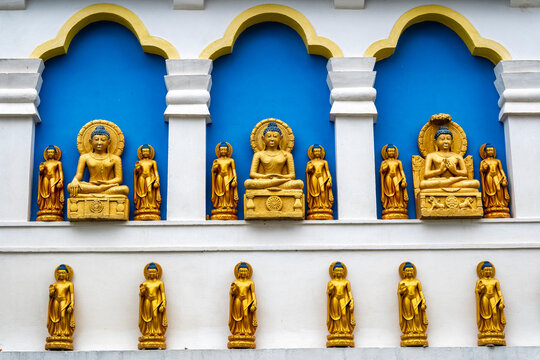 Chokling Monastery, Bir, Himachal Pradesh, India. Golden statues of Buddhas and bodhisattvas arranged along a white wall with blue and gold arches.