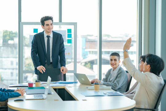 Business leader listens as colleague raises hand during presentation. Team shares questions, reviews analytics, encourages participation, collaboration, communication, stronger business decisions.