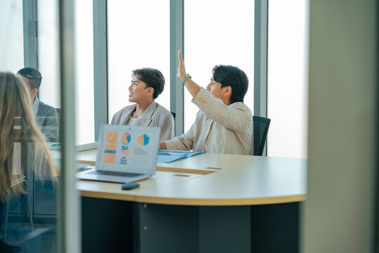 Business professionals attend conference while colleague raises hand to join discussion. Team reviews presentation, shares opinions, improves communication, engagement, teamwork, productivity.