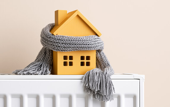 House model wrapped in scarf on radiator indoors, space for text. Winter heating efficiency