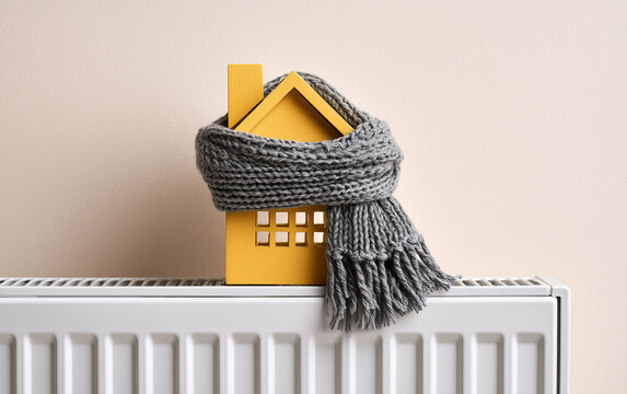 House model wrapped in scarf on radiator indoors, space for text. Winter heating efficiency