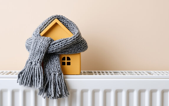 House model wrapped in scarf on radiator indoors, space for text. Winter heating efficiency