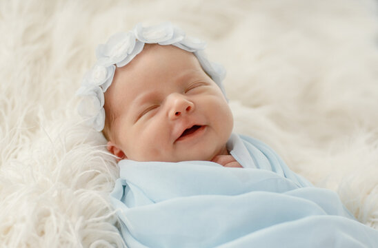 Newborn baby rests peacefully on white furry blanket. Infant wears light blue swaddle and floral bonnet, smiling gently.