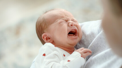 Small newborn baby wears white heart-patterned onesie. Cries loudly, eyes squeezed shut, mouth open.