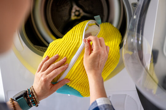 Person's hands place yellow zippered laundry bag into washing machine drum. Completing daily chores.