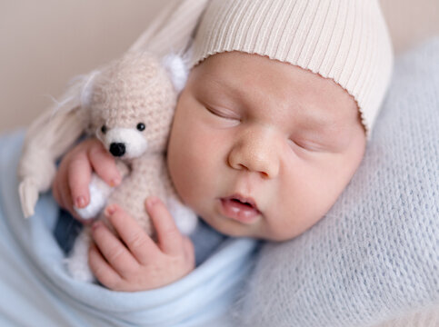 Newborn baby sleeps peacefully, wrapped in blue blanket. Wearing hat, baby holds crochet teddy bear. Professional newborn photoshoot.