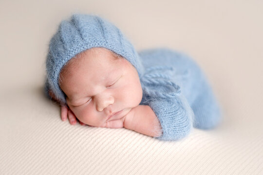 Newborn baby sleeps peacefully on soft beige blanket. Wears blue knitted bonnet and romper. Professional newborn photoshoot.