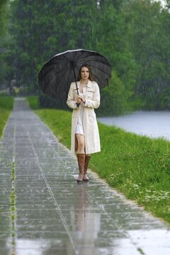 Woman walks along wet path next to river while holding black umbrella. Rain falls heavily around her. Green grass and trees are visible in background. Scene shows typical summer weather