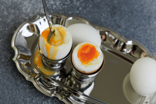 Soft boiled eggs served in silver egg cups on a metallic tray with a spoon next to them at breakfast time