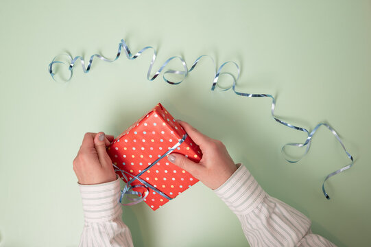Gift unwrapping: Hands pull the ribbon from a wrapped gift. Top view of a red polka dot present on a mint green background