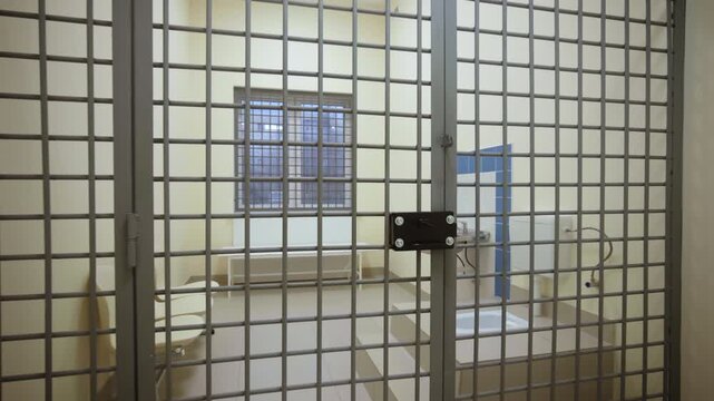 View through metal bars reveals empty holding cell with window and small sink. Strict minimalist interior creates isolating atmosphere