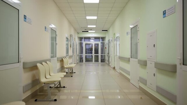 Hospital corridor shows rows of plastic chairs beside closed clinic doors. Bright ceiling lighting fills modern institutional hallway setup