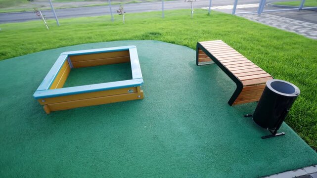 Empty playground shows sandbox wooden benches and green safety coating. Bright daylight over fresh lawn creates pleasant calm atmosphere