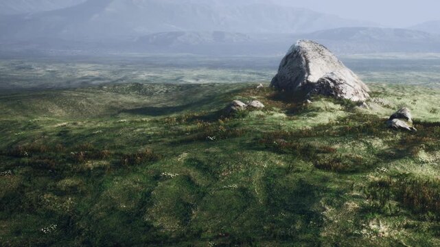 A view of a vast landscape with grass and rocks. The scene shows rolling hills in the background and a rocky formation in the foreground. It is bright and the sky is clear.