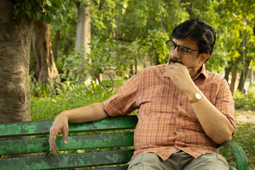 Fototapeta premium Thoughtful Indian mid aged man sitting alone on a park bench with a serious and worried expression. Concept of stress, anxiety, loneliness, mental health, retirement challenges, financial concerns.