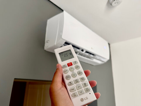 Hand holding a white LG air conditioner remote control displaying 23 degrees Celsius, pointed toward a modern wall-mounted split AC unit in a bright indoor room