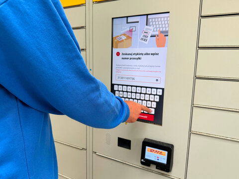 Person wearing blue jacket typing package tracking number on touchscreen parcel locker terminal for convenient self-service delivery pickup