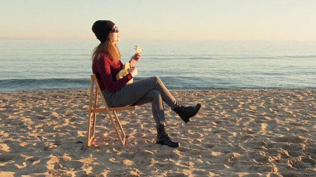Creative young musician sitting on a wooden chair on the sandy shore, playing a small ukulele and enjoying the beautiful sunset over the calm sea, expressing her freedom and creativity