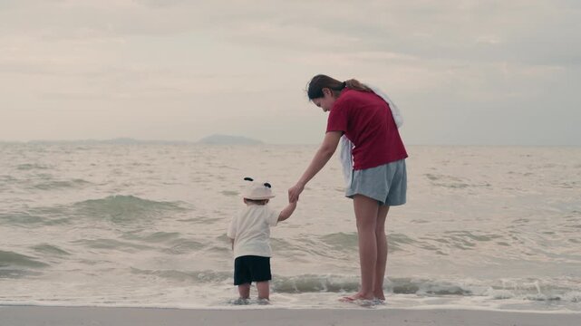 Asian mother holding hands with her baby standing on beach near ocean waves.	