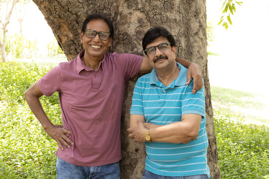 Two happy Indian senior citizens standing together with folded arms in a public park, smiling at the camera. Portrait of elderly male friends enjoying companionship, active retirement, friendship.