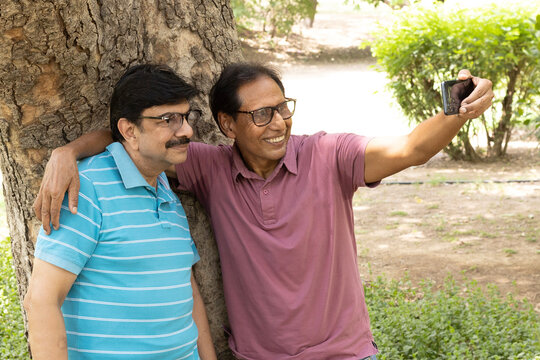 Two happy Indian senior male friends taking a selfie with a smartphone while enjoying time together in a green park. Concept of friendship, active aging, digital lifestyle, social connection.