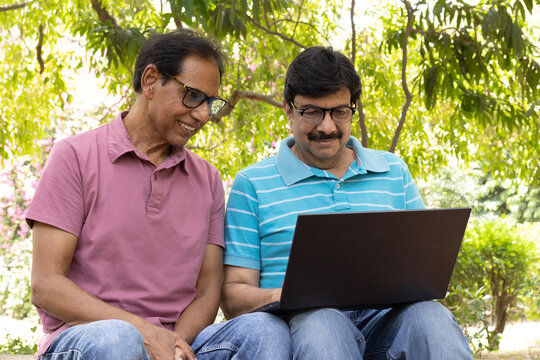 Two Indian senior citizens using a laptop together while sitting in a public park. Elderly men learning digital technology, browsing online, and discussing information, highlighting digital literacy, 