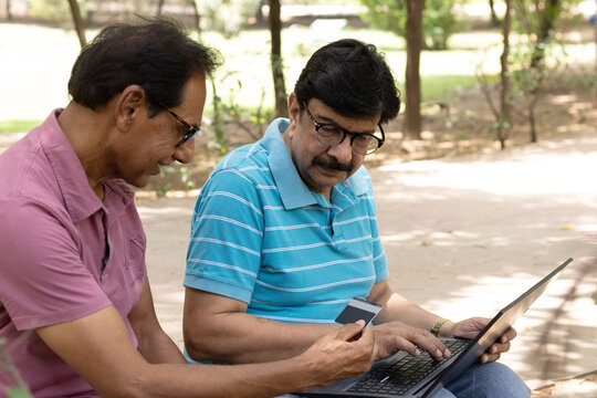 Two Indian senior citizens using a laptop together while sitting in a public park. Elderly men learning digital technology, browsing online, and discussing information, highlighting digital literacy, 