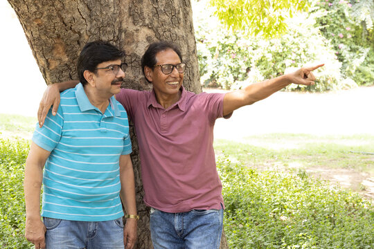 Two Indian senior male friends standing together in a park, smiling and pointing with their fingers toward copy space. Concept of friendship, active aging, outdoor lifestyle, retirement, communication