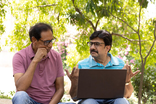 Two Indian senior citizens using a laptop together while sitting in a public park. Elderly men learning digital technology, browsing online, and discussing information, highlighting digital literacy, 