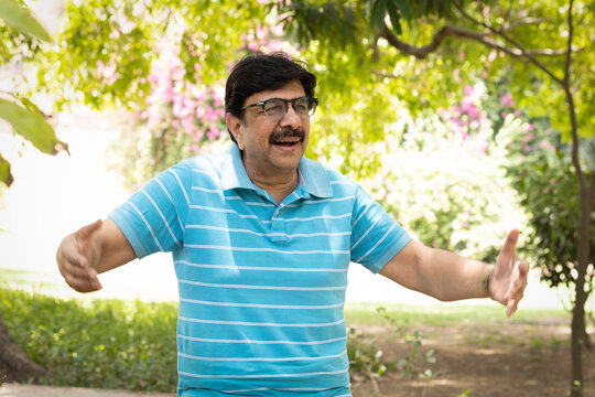 Happy Indian senior citizen standing with open arms in a lush green public park, enjoying nature and an active retirement lifestyle. Smiling elderly man wearing eyeglasses, expressing freedom.