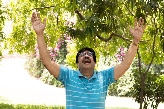 Happy Indian senior citizen standing with open arms in a lush green public park, enjoying nature and an active retirement lifestyle. Smiling elderly man wearing eyeglasses, expressing freedom.