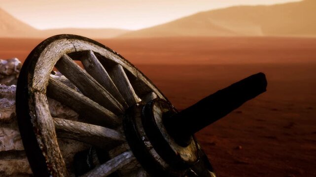 A wooden wheel leans against a stone wall in a desert. The ground is dry and red. Mountains can be seen in the background under a bright sky. It looks like a quiet place.
