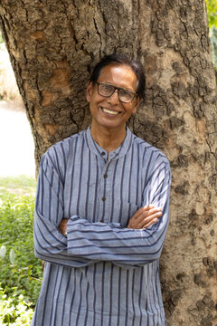Happy Indian senior citizen smiling confidently with crossed arms in a lush green park. Portrait of a cheerful elderly man wearing eyeglasses, enjoying an active and healthy retirement lifestyle in a 