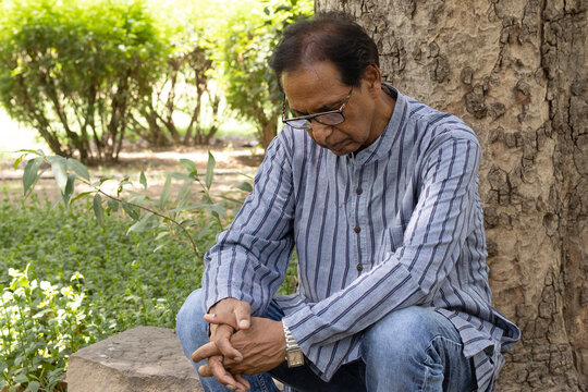 Sad Indian senior citizen sitting alone in a public park, looking thoughtful and emotional. Mature man wearing eyeglasses and traditional casual clothing, expressing loneliness, depression, aging.