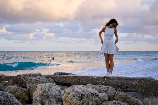 A woman in white stands on a rocky jetty with the ocean stretching behind her