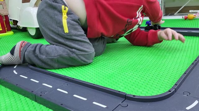 Child playing with toy car on plastic road track. Little boy sitting on green mat inside playroom. Development of imagination and creative thinking during early childhood education phase.