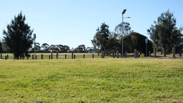 An open grassy field in a suburban public park in Hoppers Crossing, Melbourne, Australia.&nbsp;Timber bollards line the perimeter of the lawn,&nbsp;with solar street lamps&nbsp;under a clear blue sky.
