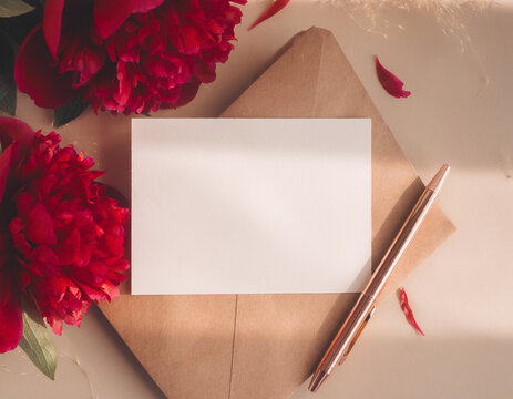 Beautiful blank card mockup with red peonies flowers and brown kraft envelope on beige background for wedding invitation or romantic note