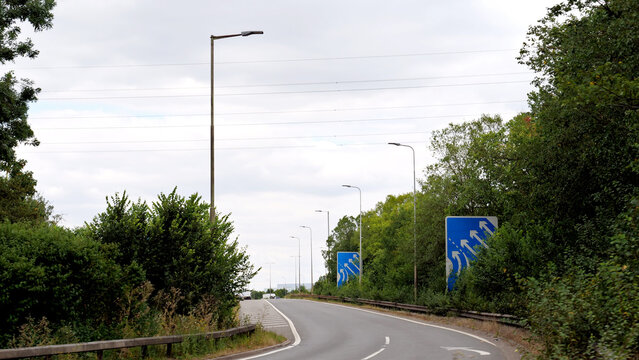 Road, highway, street, direction, sign, tree, asphalt curved road with traffic signs and street lights framed by green trees under overcast sky