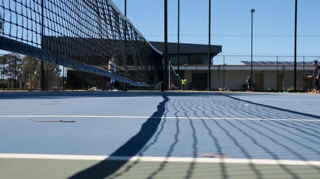 Tennis net and its shadow stretching across the blue court surface while blurred figures play in the background. A relaxed community sports venue, typical of a local tennis club in suburban Australia