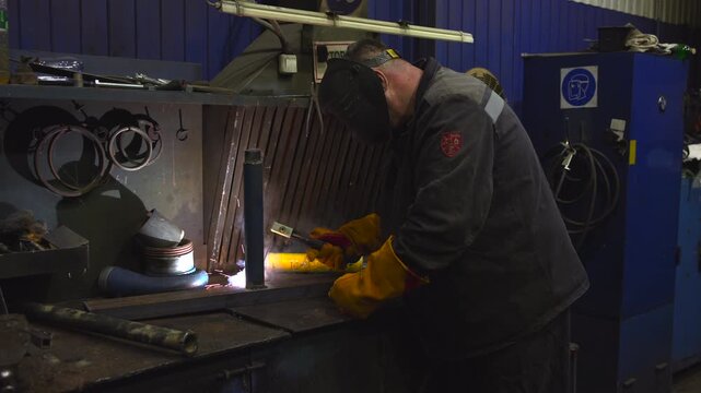 Welder performing bench welding on vertical metal post in dim industrial workshop with sparks, bright arc light, heavy gloves, helmet, steel tools, and smoky gritty atmosphere
